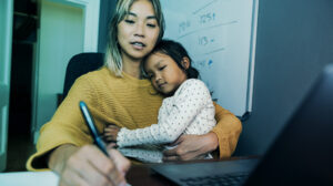 Mother holding young child while writing and working on her laptop in a home office. (Photo)
