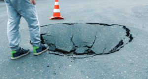 The legs of a child in grey sweatpants at the edge of a sink hole in asphalt with a traffic cone in the background.