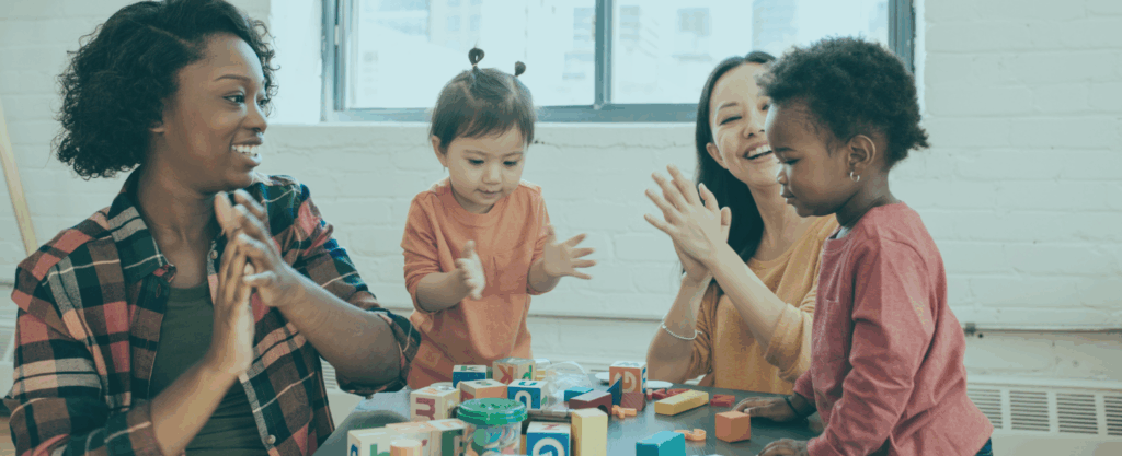 Two adults and two toddlers are sitting at a table, clapping and smiling while playing with colorful alphabet blocks in a bright, cheerful room.
