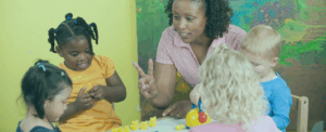 A teacher in a pink shirt is showing two fingers while speaking to a group of preschoolers engaged in play with yellow toy animals at a classroom table.