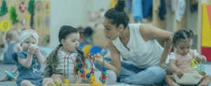 A caregiver is sitting on the floor with three young children in a playroom. One child holds a ball, another explores a toy with beads, and the caregiver is talking animatedly with the group.