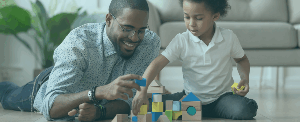 A father and his young son are lying on the floor playing together with colorful wooden blocks and shapes in a living room setting.