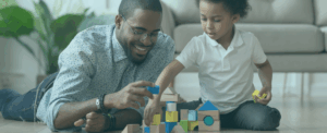A father and his young son are lying on the floor playing together with colorful wooden blocks and shapes in a living room setting.