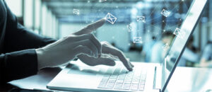 A person's hands typing on a laptop with white email icons floating around in the background.