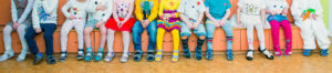 A line of children wearing colourful clothing and sitting on a bench in a classroom setting.